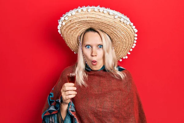 美しいですCaucasianブロンド女性身に着けているお祝いのメキシコのポンチョを飲むTequilaショット怖がって驚きのために口を開けて驚いて 不信顔 — ストック写真