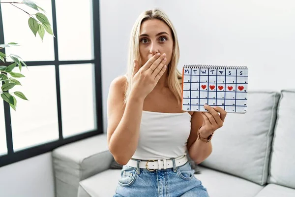 心臓のカレンダーを保持する若いブロンドの女性は間違いのために手で口をカバーショックを受けた 秘密の概念 — ストック写真