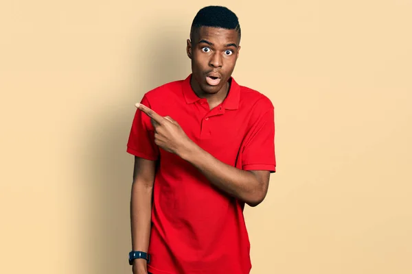 若いアフリカ系アメリカ人の男を着てカジュアルな赤いTシャツ驚きを指して指で側に 口を開けて驚いて表現 — ストック写真
