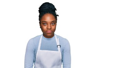 Yüzünde ciddi bir ifade olan önlük giyen Afro-Amerikan genç bir kadın. Basit ve doğal bir şekilde kameraya bakıyorsun.. 