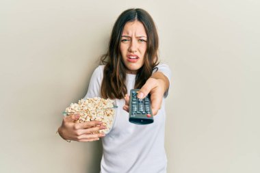 Genç esmer kadın, televizyon kumandası kullanarak patlamış mısır yiyor ve kafası karışık bir ifade kullanıyor. şüphe kavramı. 