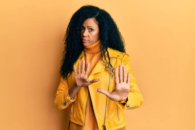 Orta yaşlı Afro-Amerikalı kadın yün kazak ve deri ceket giyiyor. Avuçlarını kaldırıyor, reddediyor ve iğrenç bir ifadeyle reddediyor. Dur ve yasakla. 