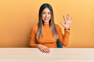 Latin kökenli genç bir kadın, gündelik giysiler içinde masada oturuyor ve beş numaralı parmaklarını göstererek gülümsüyor ve mutlu.. 