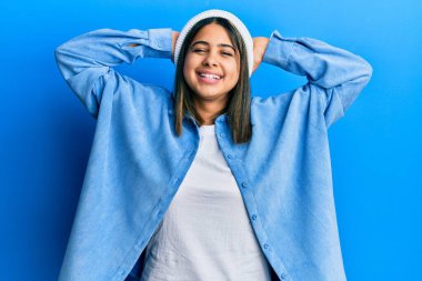 Tatlı yün şapkalı genç Latin kadın rahatlıyor ve esniyor, kolları ve elleri başının ve boynunun arkasında mutlu bir şekilde gülümsüyor. 