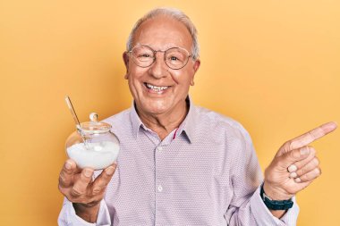 Gri saçlı, şeker dolu kaseli yaşlı adam gülümsüyor mutlu bir şekilde elini ve parmağını yana doğru gösteriyor. 