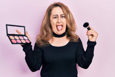 Orta yaşlı beyaz bir kadın elinde makyaj fırçası ve utangaç bir dille komik bir ifadeyle mutlu bir şekilde dışarı çıkıyor.. 