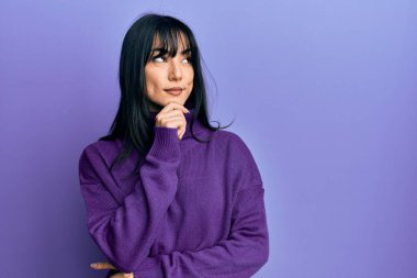 Kâküllü, boğazlı kazak giyen, eli çenesinde soru soran genç esmer kadın, düşünceli bir ifade. Düşünceli bir yüzle gülümsüyor. şüphe kavramı. 