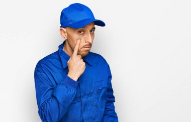 Sakallı, inşaatçı tulumu giymiş kel bir adam. Gözünü dikip jestini izliyor. Şüpheli bir ifade. 