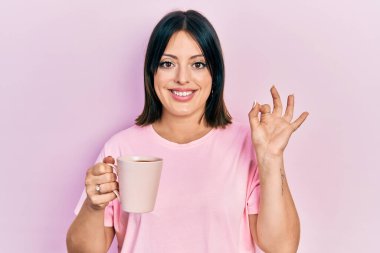 Bir fincan kahve içen genç İspanyol kadın parmaklarıyla imza atıyor, gülümseyerek mükemmel bir sembol oluşturuyor. 