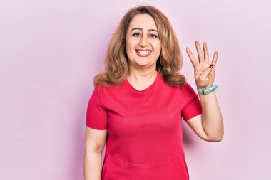 Orta yaşlı beyaz kadın, günlük kıyafetler giyip dört numaralı parmaklarıyla işaret ederken kendinden emin ve mutlu bir şekilde gülümsüyor.. 