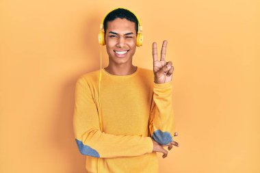 Genç Afro-Amerikalı adam müzik dinliyor kulaklıkla gülümsüyor kameraya göz kırparak parmaklarıyla zafer işareti yapıyor. İki numara.. 