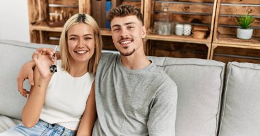 Genç, beyaz bir çift gülümsüyor. Kanepede yeni evlerinin anahtarını tutuyorlar..