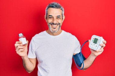 Yakışıklı, orta yaşlı, gri saçlı, kan basıncı monitörü kullanan yüzünde mutlu ve havalı bir gülümsemeyle gülümseyen bir adam. diş gösterme. 