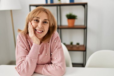 Orta yaşlı, sarışın, gülümseyen mutlu, rahat bir şekilde evdeki masada oturuyor..