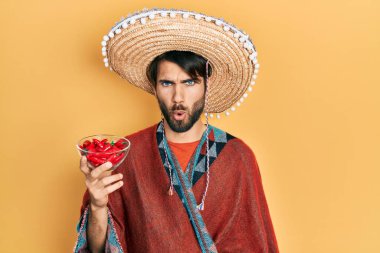 Meksikalı şapkalı genç İspanyol adam elinde kırmızı biber tutarken korkmuş ve ağzı açık şaşkınlık içinde şaşırmış, inançsız yüz. 