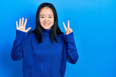 Genç Çinli kız gündelik giysiler giyer ve yedi numaralı parmaklarıyla işaret ederken kendinden emin ve mutlu gülümserdi.. 