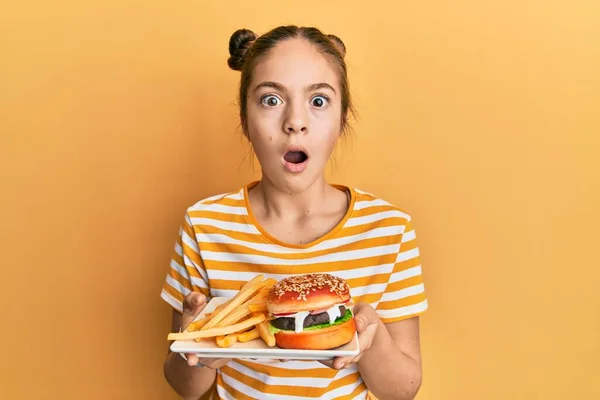 美しいブルネットの女の子はフライでおいしい古典的なバーガーを食べている恐怖と驚きと驚きの表情 恐怖と興奮した顔でショックを受けました — ストック写真