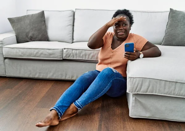 自宅の床に座っているスマートフォンを使用して若いアフリカの女性は非常に幸せと笑みを浮かべて遠くに手を頭の上に見て 検索の概念 — ストック写真