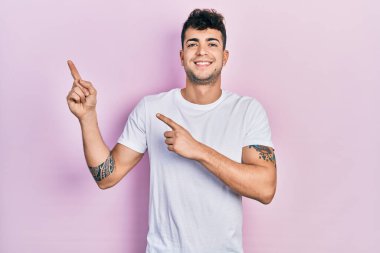 Gündelik beyaz tişört giyen genç İspanyol adam gülümseyip kameraya bakıyor ve iki eliyle ve parmaklarıyla kenarı işaret ediyor.. 