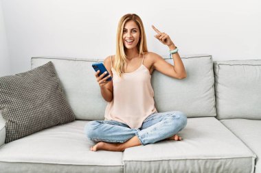 Sarışın, güzel, genç bir kadın. Kanepede oturuyor. Akıllı telefon tebessümü kullanıyor. 