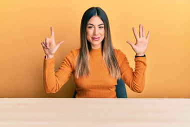 Genç İspanyol kadın, gündelik giysiler içinde masada oturuyor ve yedi numaralı parmaklarıyla işaret ederken kendinden emin ve mutlu gülümsüyor.. 