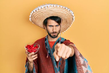 Meksika şapkalı genç İspanyol adam elinde kırmızı biber ile kamerayı işaret ediyor ve sen kendine güvenen ciddi görünüyorsun. 