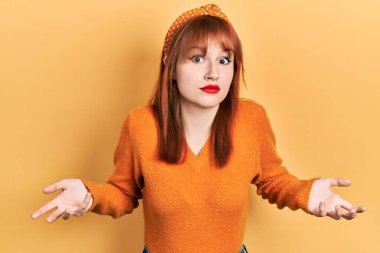 Kızıl saçlı, turuncu süveter giyen, elleri ve kolları havada, kafası karışmış genç bir kadın. şüphe kavramı. 