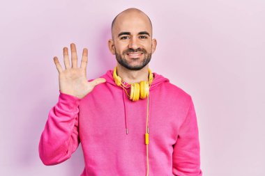 Spor kıyafetleri giyen, kulaklık takan genç kel adam beş numarayı gösterip işaret ederken kendinden emin ve mutlu bir şekilde gülümsüyor.. 