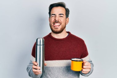 Sakallı yakışıklı beyaz bir adam bir fincan kahve içiyor termosu var, dilini dışarı çıkarıyor. Komik bir ifadeyle.. 