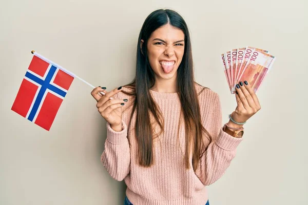 年轻的惊慌失措的女人举着诺威国旗和克朗钞票 带着滑稽的表情伸出舌头 高兴极了 — 图库照片