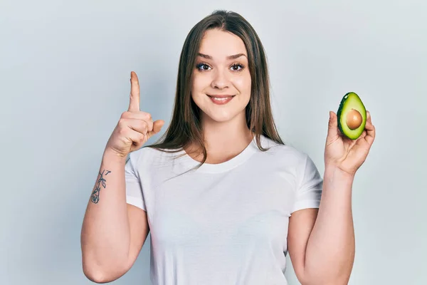 Young Hispanic Girl Holding Middle Avocado Smiling Idea Question Pointing — ストック写真