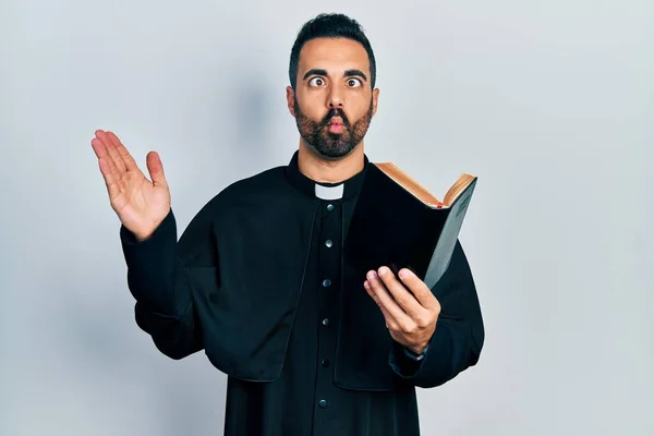 英俊而惊慌失措的牧师 留着大胡子 手里拿着圣经和基督教十字架 用嘴和眼神做鱼脸 疯狂而滑稽 — 图库照片