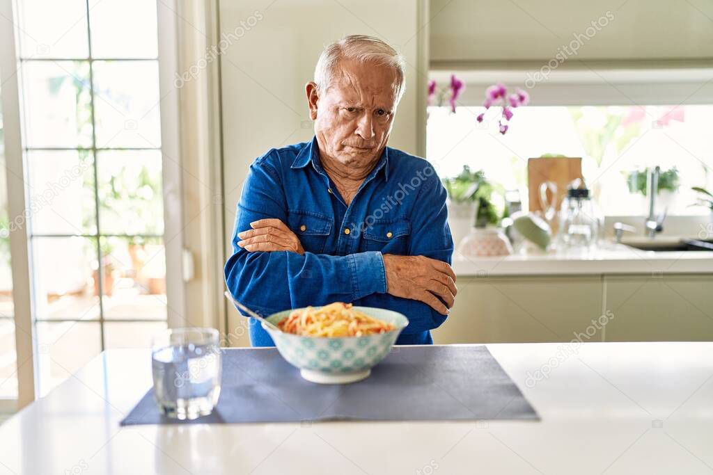 Senior man with grey hair eating pasta spaghetti at home skeptic and nervous, disapproving expression on face with crossed arms. negative person. 