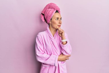 Duş havlusu şapkası takan ve eli çenesinde bornoz giyen genç sarışın kadın soru sormayı düşünüyor, düşünceli bir ifade. Gülümseyen ve düşünceli bir yüz. şüphe kavramı. 