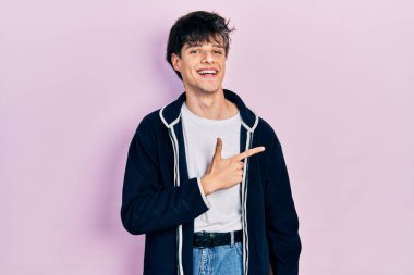 Yakışıklı hippi genç adam, günlük beyaz tişört ve sweatshirt giyiyor. Gülümsüyor, eli ve parmağı yana doğru uzanıyor. 