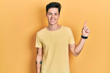 Yüzünde kocaman bir gülümseme olan sıradan sarı tişörtler giyen genç İspanyol adam kameraya el ile işaret ediyor.. 