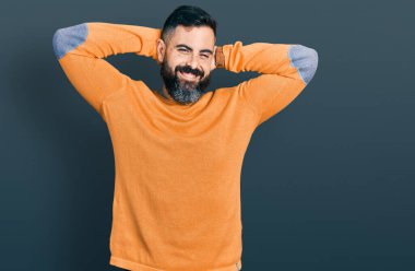 Sakallı, sıradan bir süveter giyen, kolları ve elleri başının arkasında, mutlu bir şekilde gülümseyen İspanyol bir adam. 