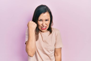 Orta yaşlı İspanyol kadın rahat kıyafetler giyiyor kızgın ve kızgın yumruğunu kaldırıyor ve öfkeyle bağırıyor. öfke ve agresif konsept. 