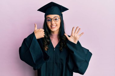 Mezuniyet şapkası ve tören cübbesi takan genç İspanyol kadın altı numarayı gösterip işaret ederken kendinden emin ve mutlu bir şekilde gülümsüyor.. 