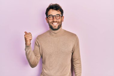 Balıkçı yaka kazak ve gülen yüzlü gözlük takan sakallı yakışıklı bir adam başparmağıyla yana bakıyor ve işaret ediyor.. 