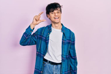 Yakışıklı hippi genç adam, günlük beyaz tişört ve klasik tişört giyiyor gülümsüyor ve kendine güvenen el hareketleriyle küçük beden işareti yapıyor, parmaklarıyla bakıyor ve kameraya bakıyor. ölçüm kavramı. 