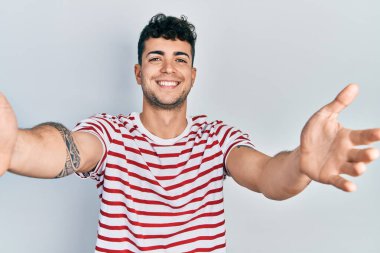 Gündelik giysiler giymiş genç İspanyol adam kameraya bakıp sarılmak için kollarını açmış gülümsüyor. Mutluluğu kucaklayan neşeli bir ifade.. 