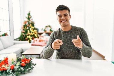 Noel ağacının yanındaki masada oturan genç İspanyol adam mutlu ve komik yüzüyle kamerayı işaret ediyor. iyi enerji ve titreşimler. 