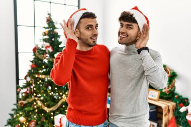 Genç eşcinsel çift Noel ağacının yanında şapkalarını takmış, kulaklarında gülümseyerek dedikoduları ve dedikoduları dinliyor. Sağırlık kavramı. 