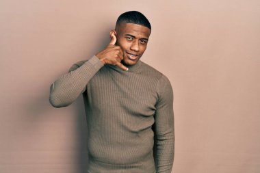 Balıkçı yaka kazak giyen genç siyahi bir adam gülümsüyor telefonla el ve parmaklarıyla jest yapıyor telefonda konuşuyor gibi. iletişim kavramları. 