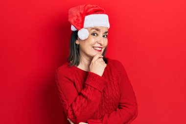 Orta yaşlı İspanyol kadın Noel şapkası takıyor kameraya kendinden emin bakıyor kollarını kavuşturmuş ve çenesini kaldırmış bir şekilde gülümsüyor. Olumlu düşünmek. 