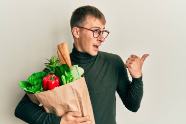Kafkasyalı genç bir adam elinde poşet, elinde alışveriş poşetleri, başparmağını yukarı kaldırmış, ağzı açık gülümsüyor. 