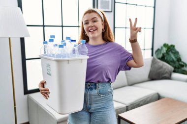 Genç kızıl saçlı kadın elinde plastik şişelerle çöp kutusuyla kameraya bakıp parmaklarını zafer işareti yaparken gösteriyor. İki numara.. 