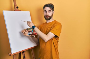 Beyaz, sakallı, palet çizen beyaz bir adam. Resim sehpası kuşkucu ve gergin duruyor. Problemden dolayı surat asıyor. negatif kişi. 