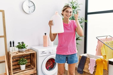 Çamaşır deterjanı şişesini tutan genç sarışın kadın bir fikirle gülümsüyor ya da bir soru işareti yapıyor. 